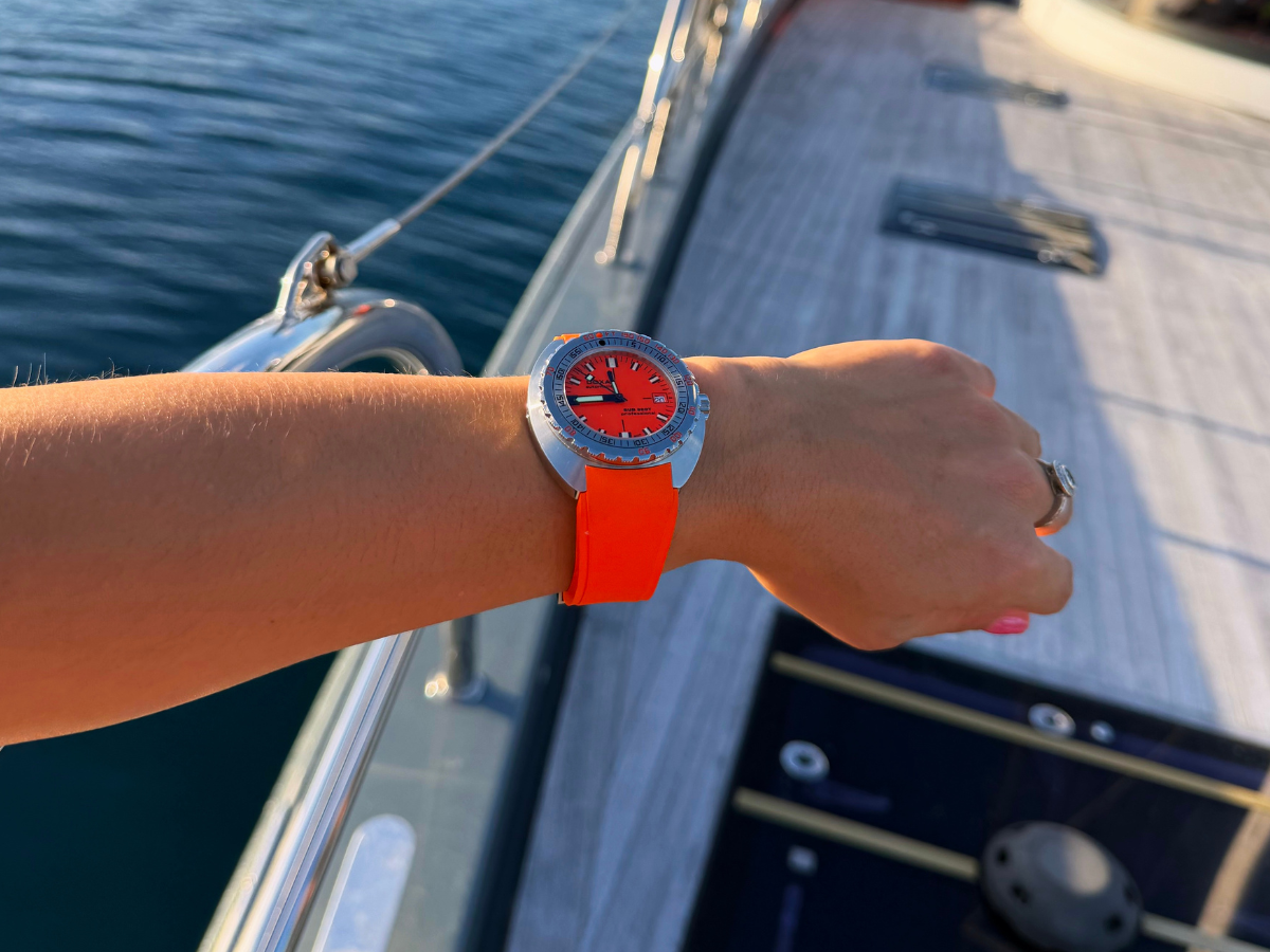 Armbanduhr mit rotem Zifferblatt, silbernem Gehäuse und orangefarbenem Kautschukband am Handgelenk, auf einem Segelboot